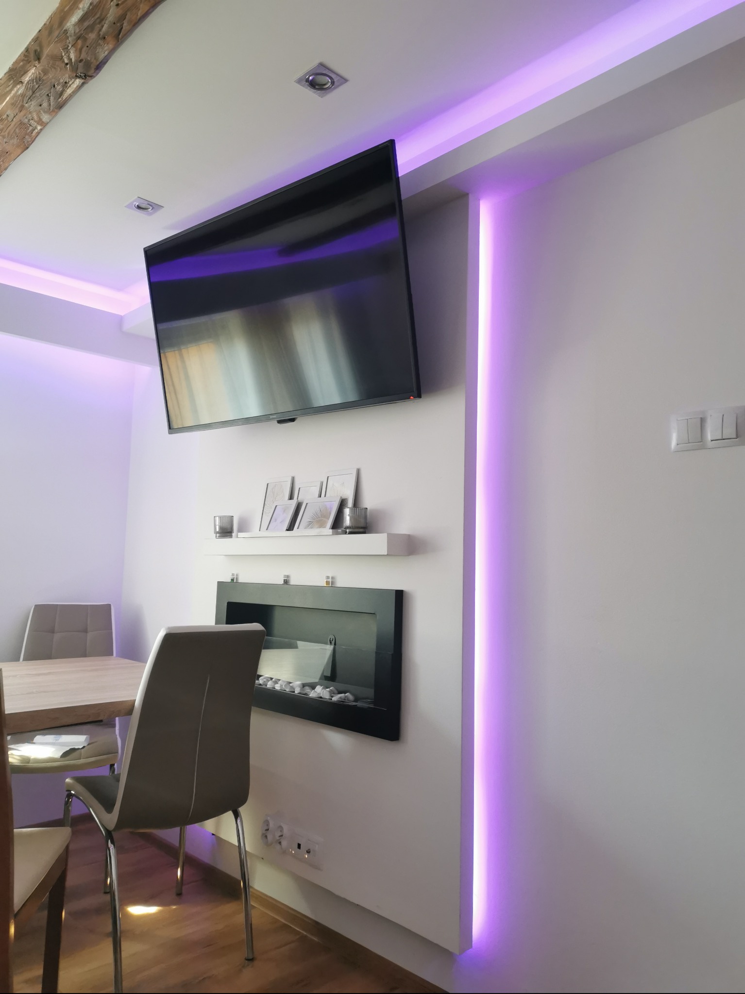 Nowoczesne wnętrze z podświetleniem LED: Ściana z telewizorem, biokominkiem i półką dekoracyjną, oświetlona fioletowym światłem. Widoczny stół z krzesłami.