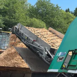 Żółta ładowarka Volvo z otwartą kabiną nabiera stertę zrębków drewna, które są transportowane podajnikiem taśmowym do zielonego kontenera na tle drzew.