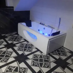 Biała wanna z hydromasażem i podświetleniem LED w nowoczesnej łazience z czarno-białą wzorzystą podłogą i marmurową ścianą.