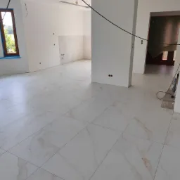 Salon w Tarnowie po ułożeniu białych, marmurkowych płytek podłogowych. Ściany pomalowane na biało, widoczne przygotowane punkty elektryczne i niedokończone prace wykończeniowe.