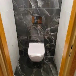 Wykończona toaleta w nowoczesnym stylu: biała miska ustępowa, czarne płytki imitujące marmur na ścianie i podłodze, chromowany przycisk spłukujący. Widok zza drewnianej futryny.