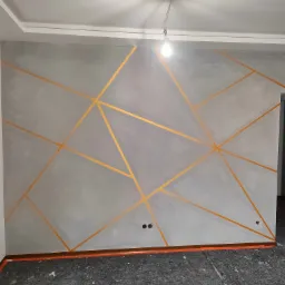 Ściana pokryta szarą farbą z geometrycznym wzorem z taśmy w kolorze złota, widoczne gniazdka elektryczne i zabezpieczona podłoga.