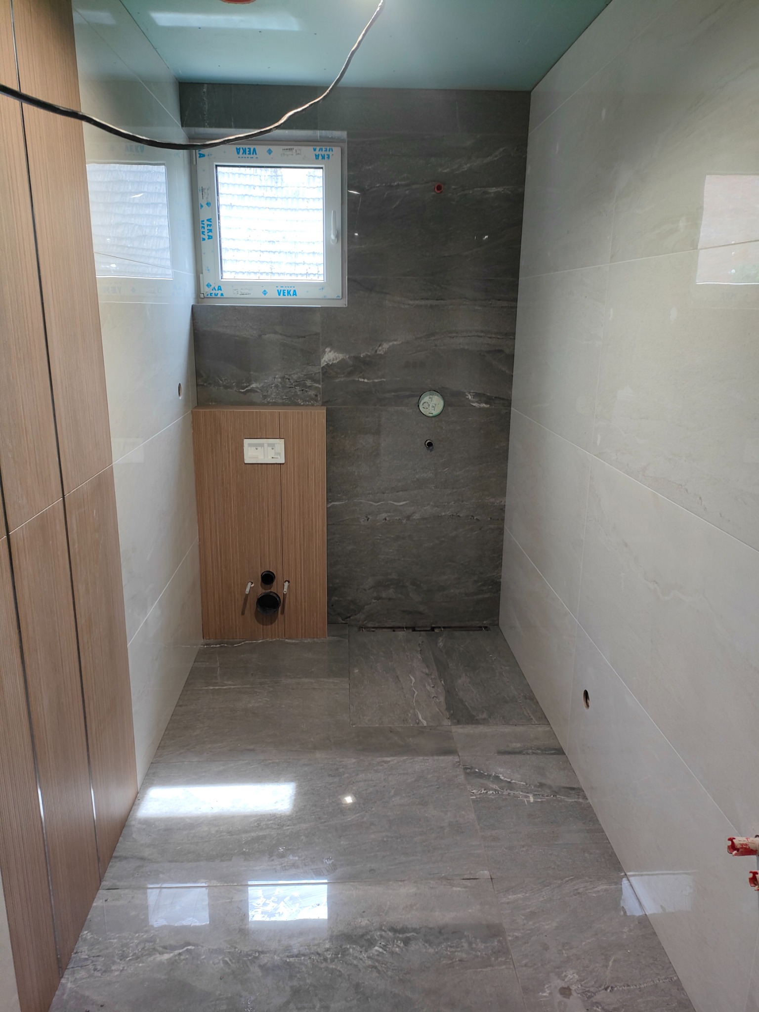 Nowoczesna łazienka w trakcie wykańczania: szare płytki podłogowe z połyskiem, drewniana zabudowa stelaża WC, białe płytki ścienne, widoczne instalacje przed podłączeniem armatury.