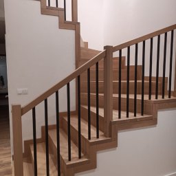 Projektowanie wnętrz - Drewniane schody z czarnymi balustradami, widok z dołu, oświetlenie górne, minimalistyczny design.