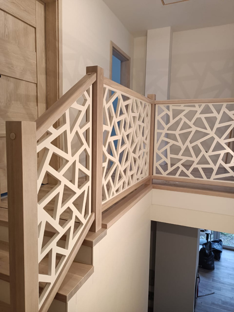 Ażurowa balustrada schodowa z drewna w geometryczny wzór, widok z góry na klatkę schodową i drzwi wejściowe.