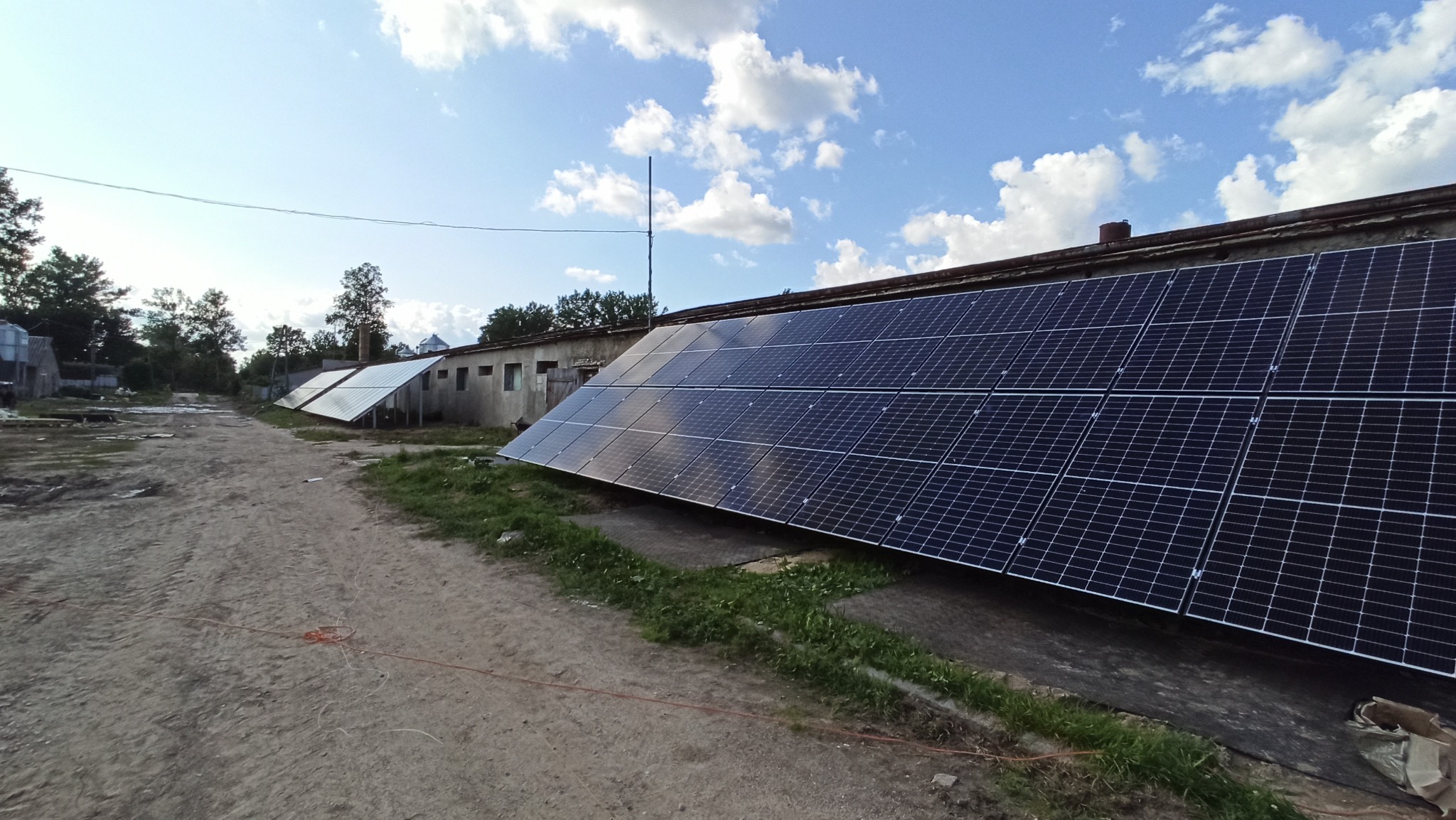 Instalacja paneli słonecznych na dachu budynku gospodarczego, widok z perspektywy gruntu, z widoczną drogą gruntową i fragmentami innych budynków w tle.