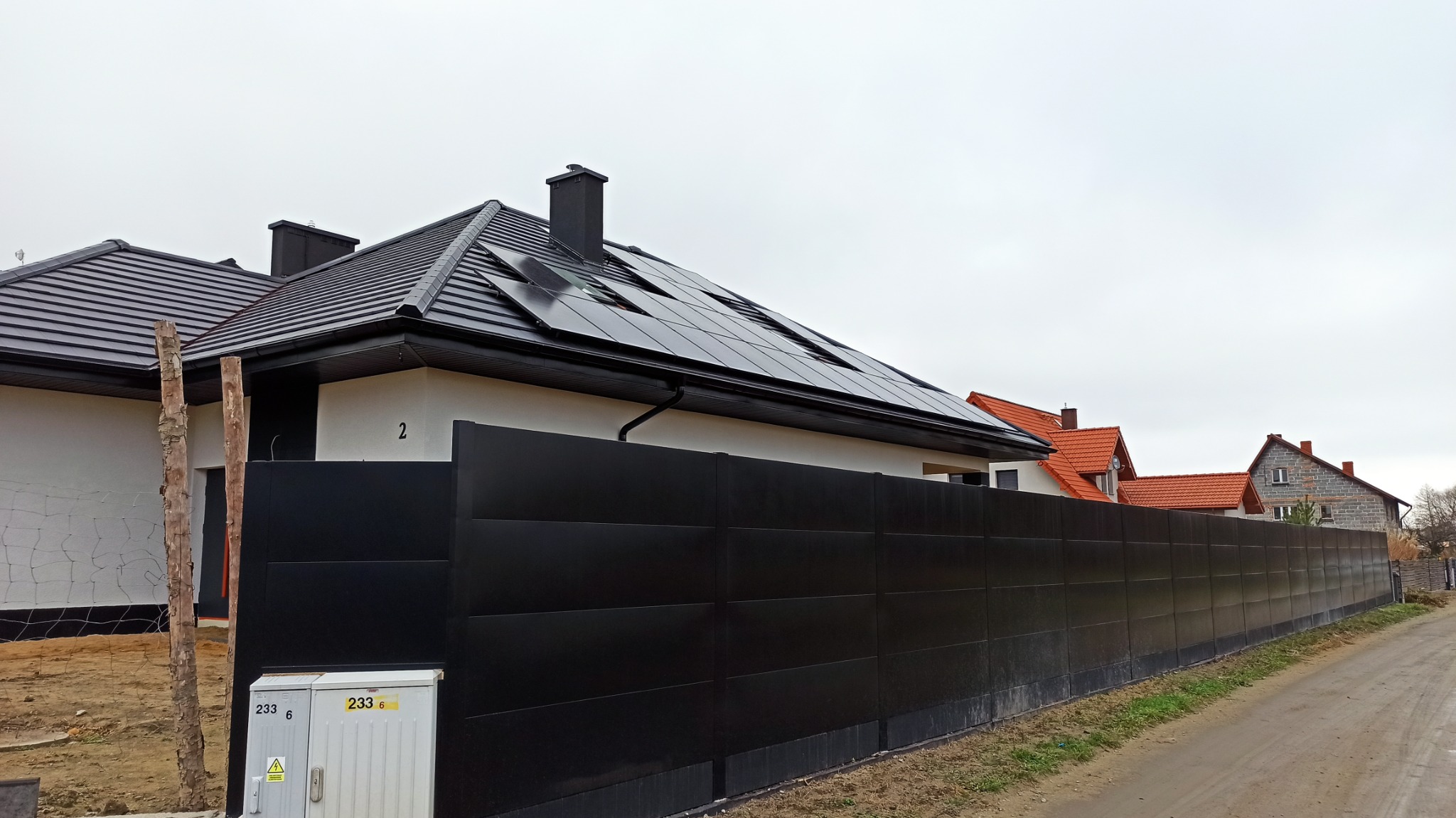 Nowoczesny dom jednorodzinny z czarnym dachem i panelami słonecznymi, otoczony wysokim, czarnym ogrodzeniem panelowym, widok z ulicy w pochmurny dzień.
