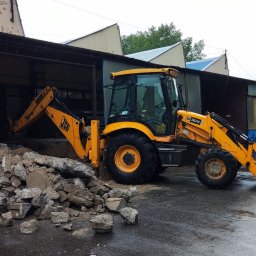 Mj Construction - Żółta koparko-ładowarka JCB 3CX na mokrym placu budowy obok sterty gruzu, w tle zadaszona hala z elementami niebieskiego dachu. Widoczne krople deszczu.