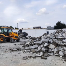 Mj Construction - Żółta koparko-ładowarka JCB 3CX podczas prac rozbiórkowych, sterta gruzu i odłamków budowlanych na placu, fragment białej ściany w tle, pochmurne niebo.