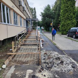 Mj Construction - Budowa podjazdu dla niepełnosprawnych przy budynku, widoczna konstrukcja szkieletowa z metalu i drewna, obok sterta gruzu, w tle idąca osoba.