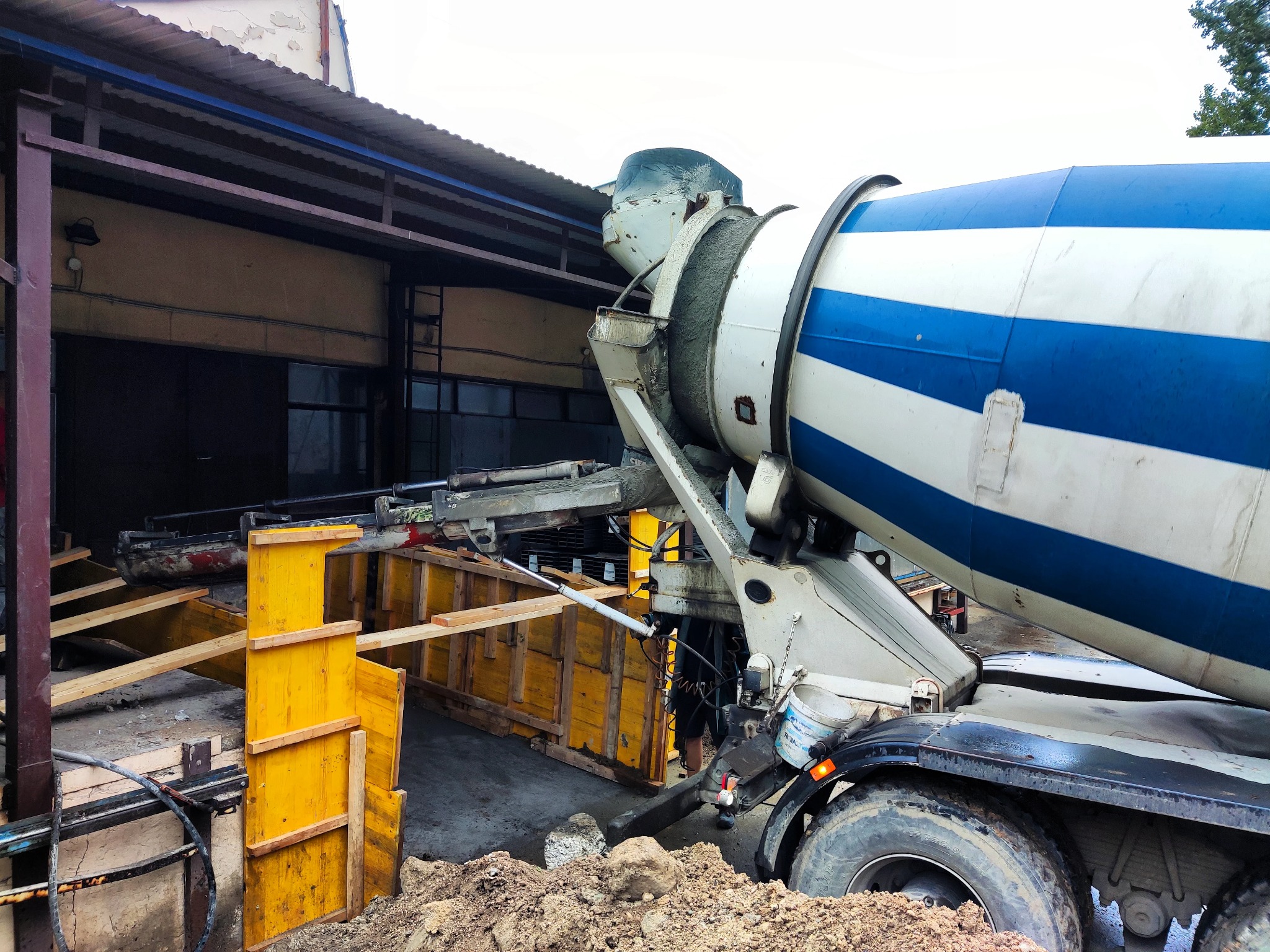 Betoniarka z niebieskimi pasami wylewa beton do żółtego szalunku fundamentowego na placu budowy.