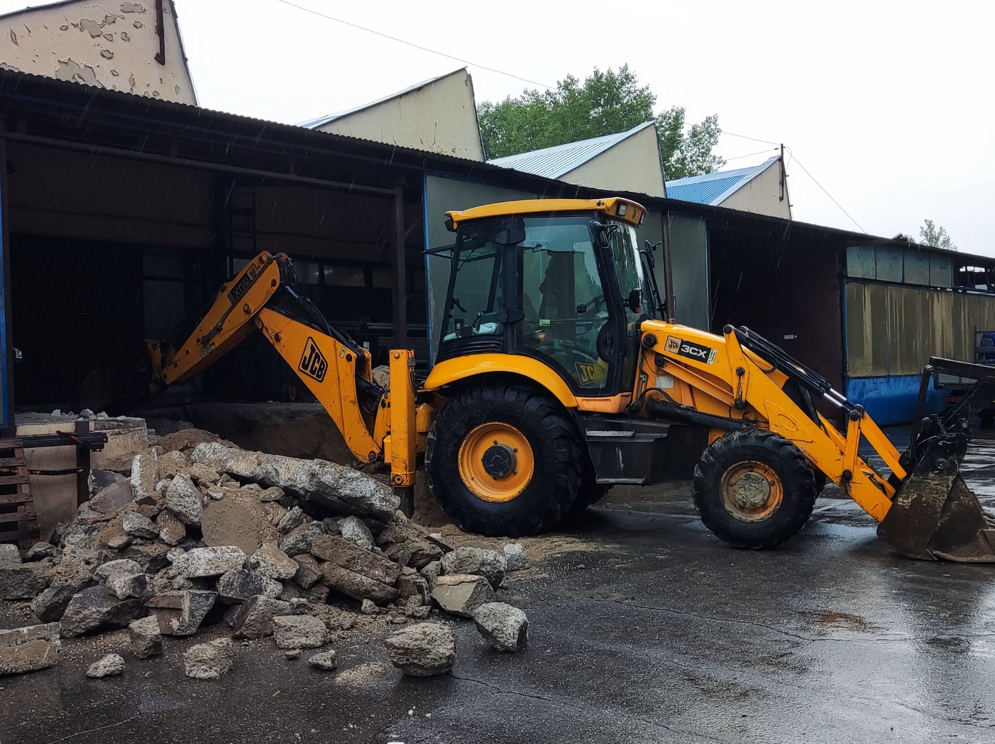 Żółta koparko-ładowarka JCB 3CX na mokrym placu budowy obok sterty gruzu, w tle zadaszona hala z elementami niebieskiego dachu. Widoczne krople deszczu.
