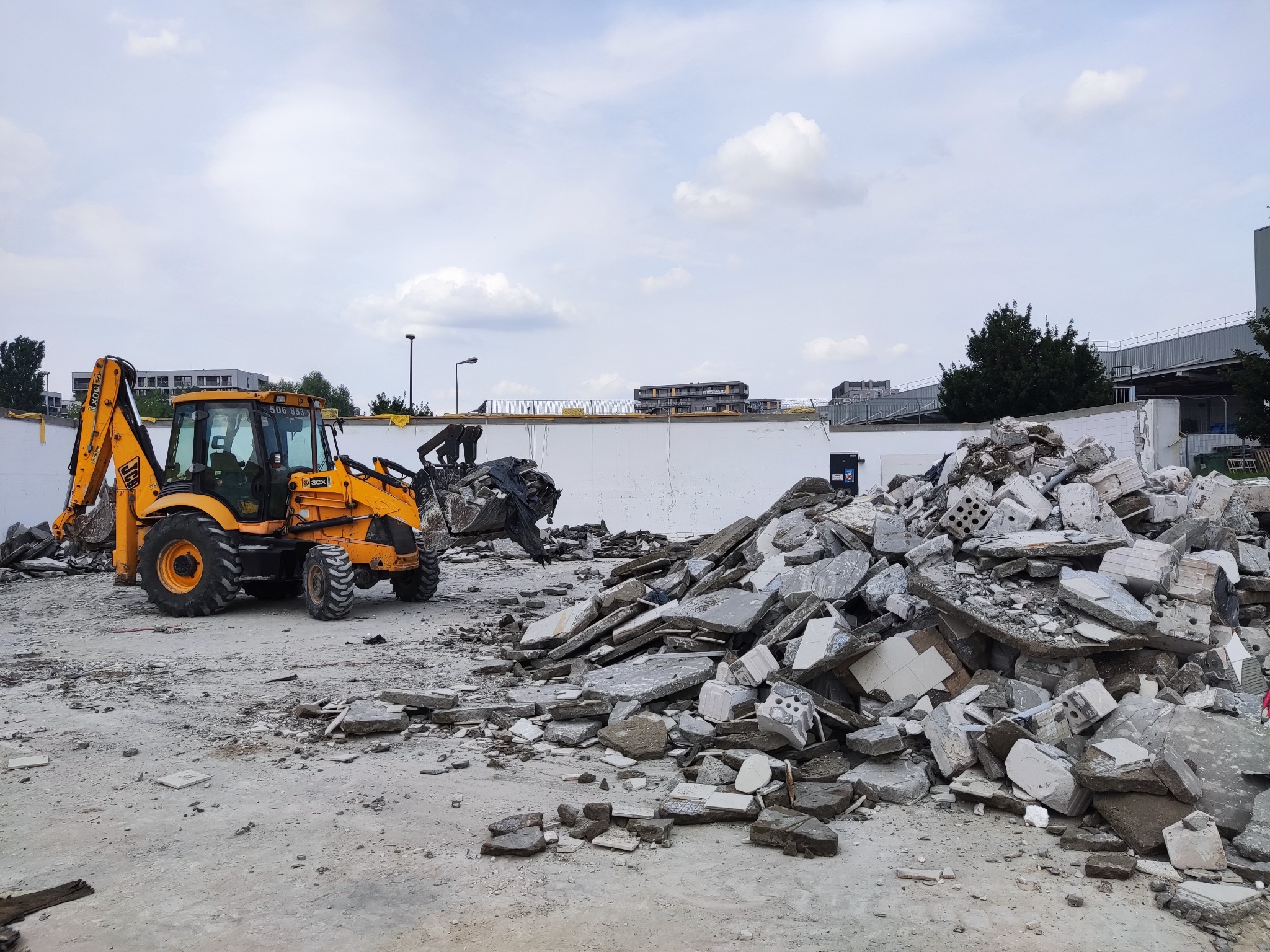 Żółta koparko-ładowarka JCB 3CX podczas prac rozbiórkowych, sterta gruzu i odłamków budowlanych na placu, fragment białej ściany w tle, pochmurne niebo.