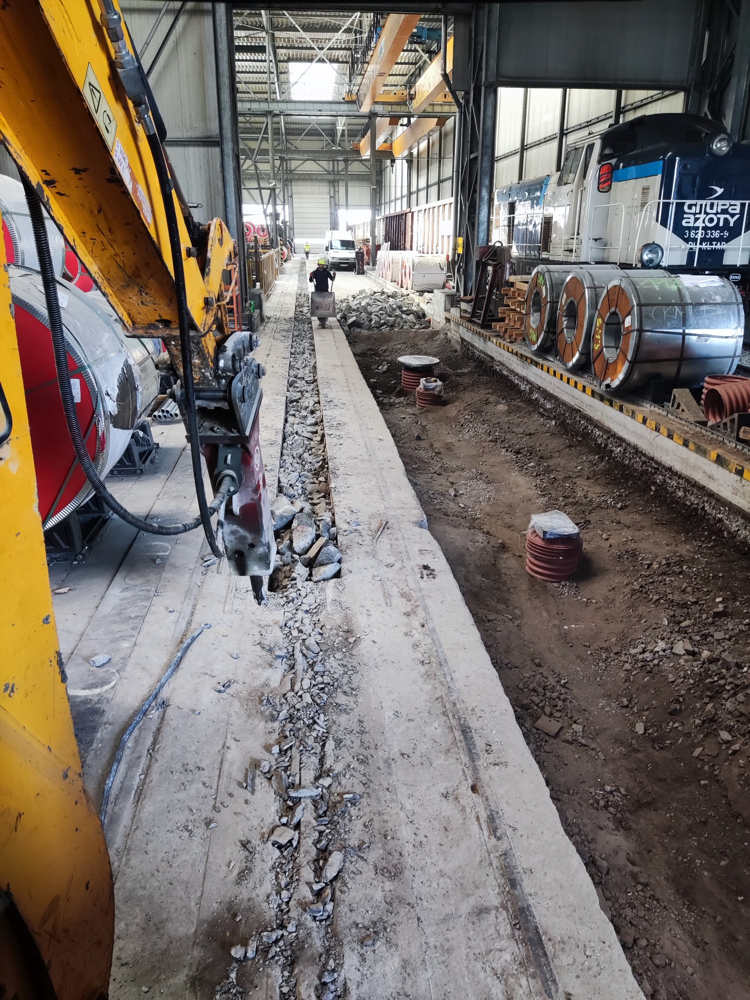 Wnętrze hali przemysłowej: robotnik z taczką w oddali, wykopany rów z gruzem, obok rury kanalizacyjne i pociąg zwojami blachy w tle, prace wyburzeniowe betonu przy użyciu młota pneumatycznego...