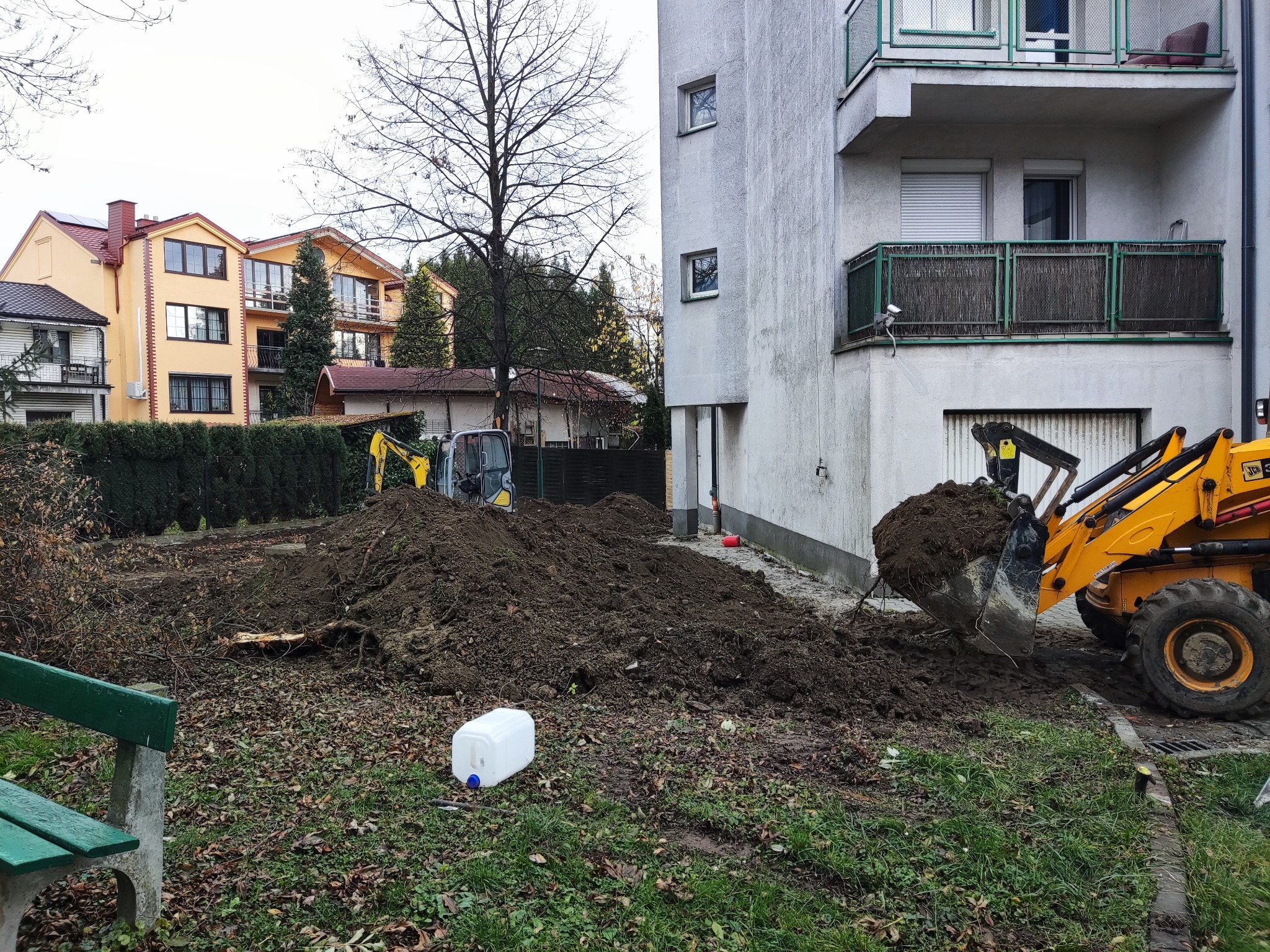 Żółta koparko-ładowarka nabiera ziemię z wykopu na posesji, obok budynku mieszkalnego z balkonami i w tle widoczne inne domy.