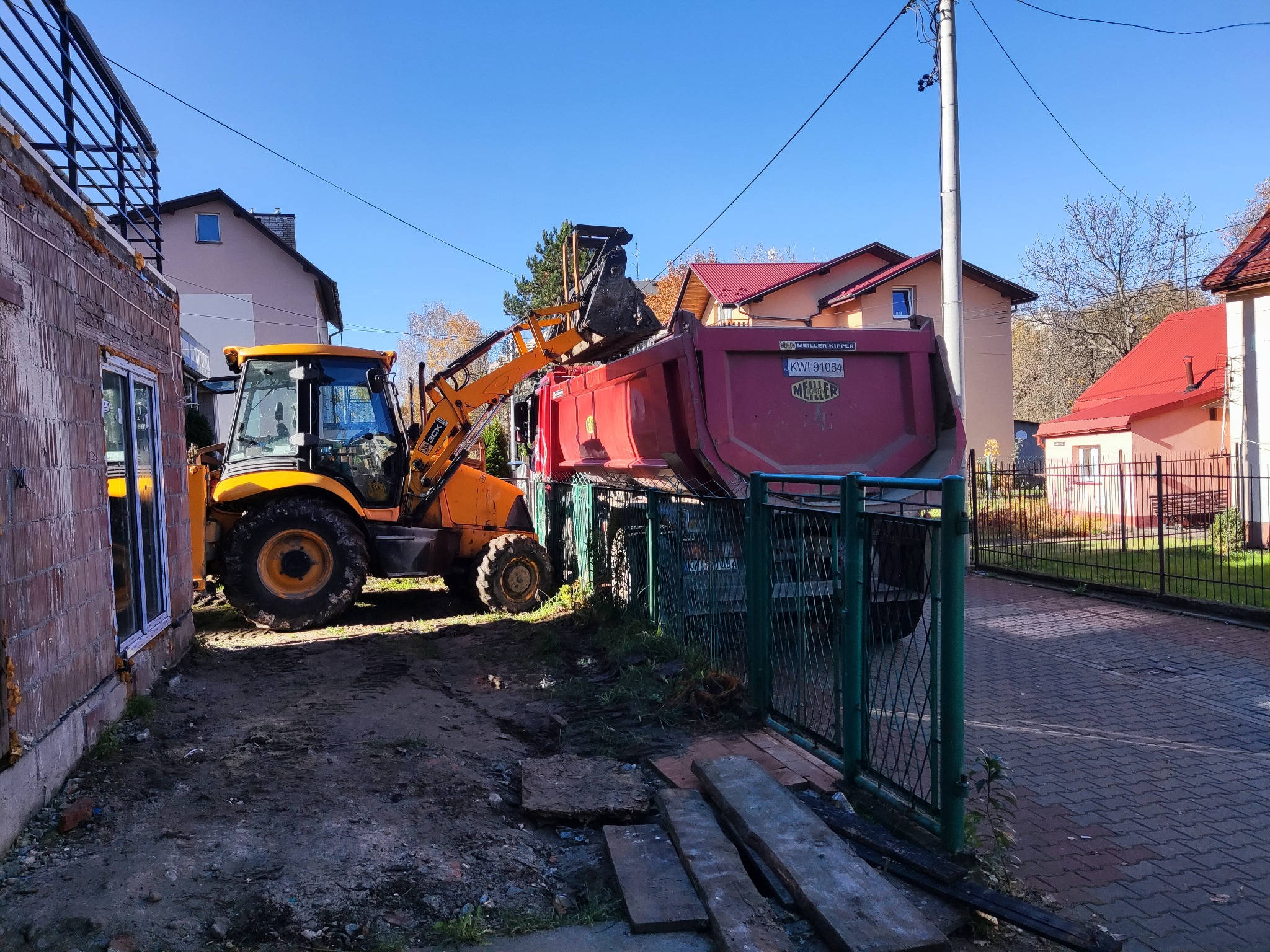 Żółta koparko-ładowarka nabiera ziemię do czerwonej wywrotki na tle budynków mieszkalnych i błękitnego nieba.