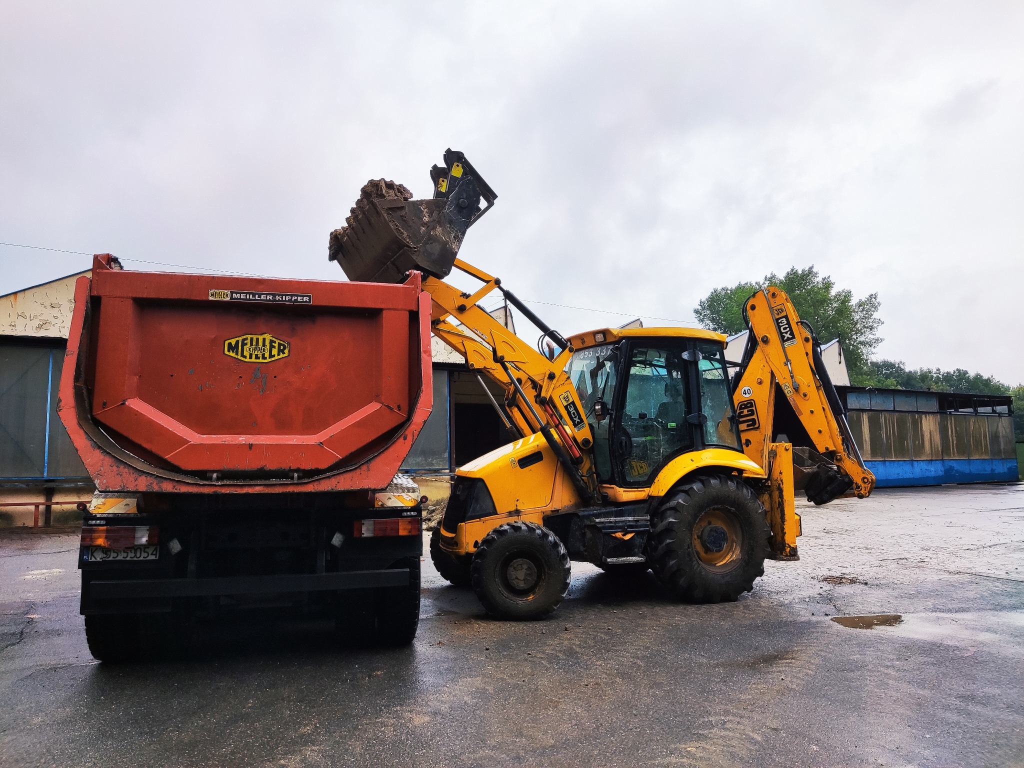 Żółta koparko-ładowarka JCB 3CX z pełną łyżką ziemi wysypuje urobek do pomarańczowej ciężarówki Meiller Kipper na mokrym placu budowy.