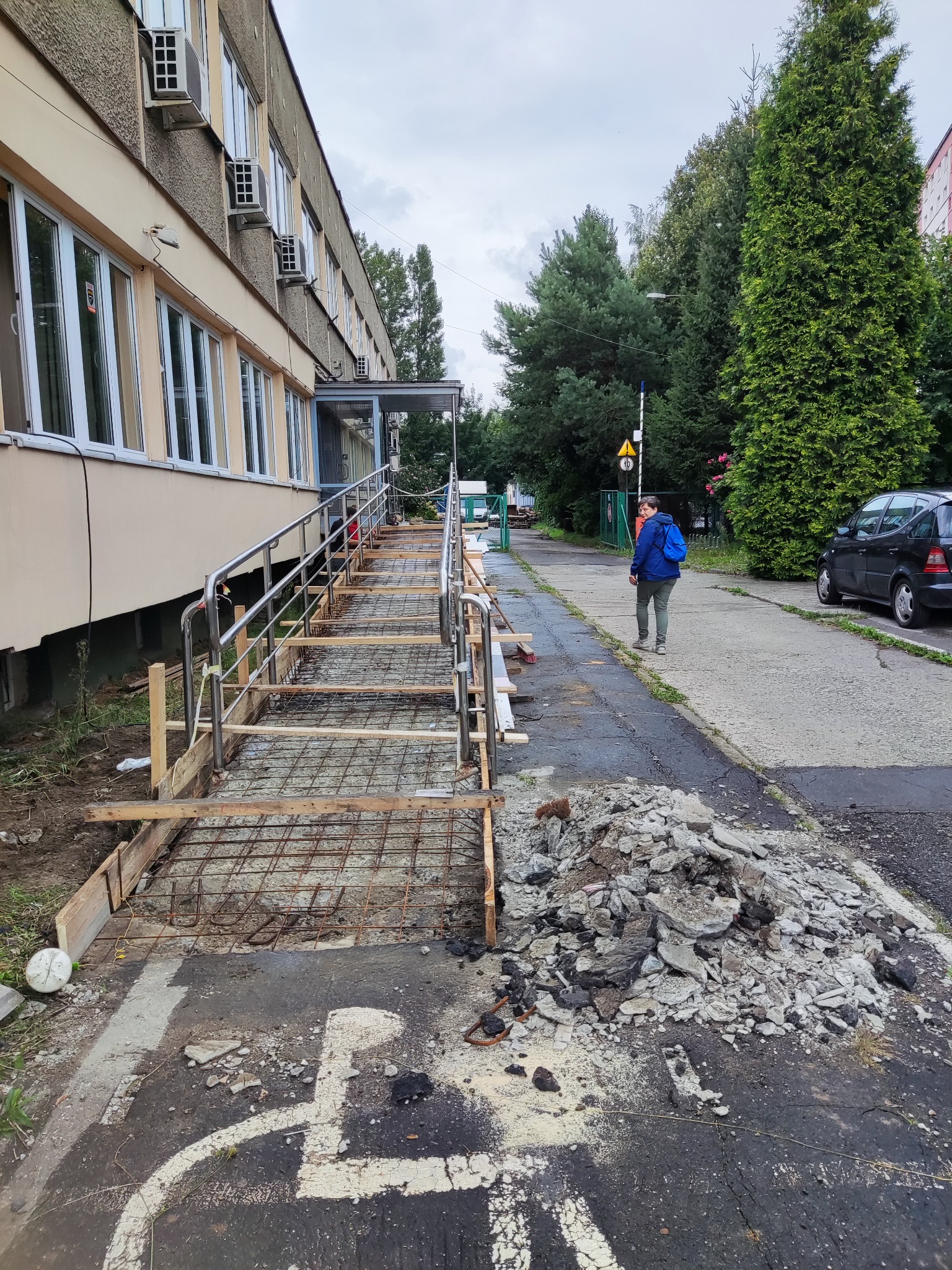 Budowa podjazdu dla niepełnosprawnych przy budynku, widoczna konstrukcja szkieletowa z metalu i drewna, obok sterta gruzu, w tle idąca osoba.