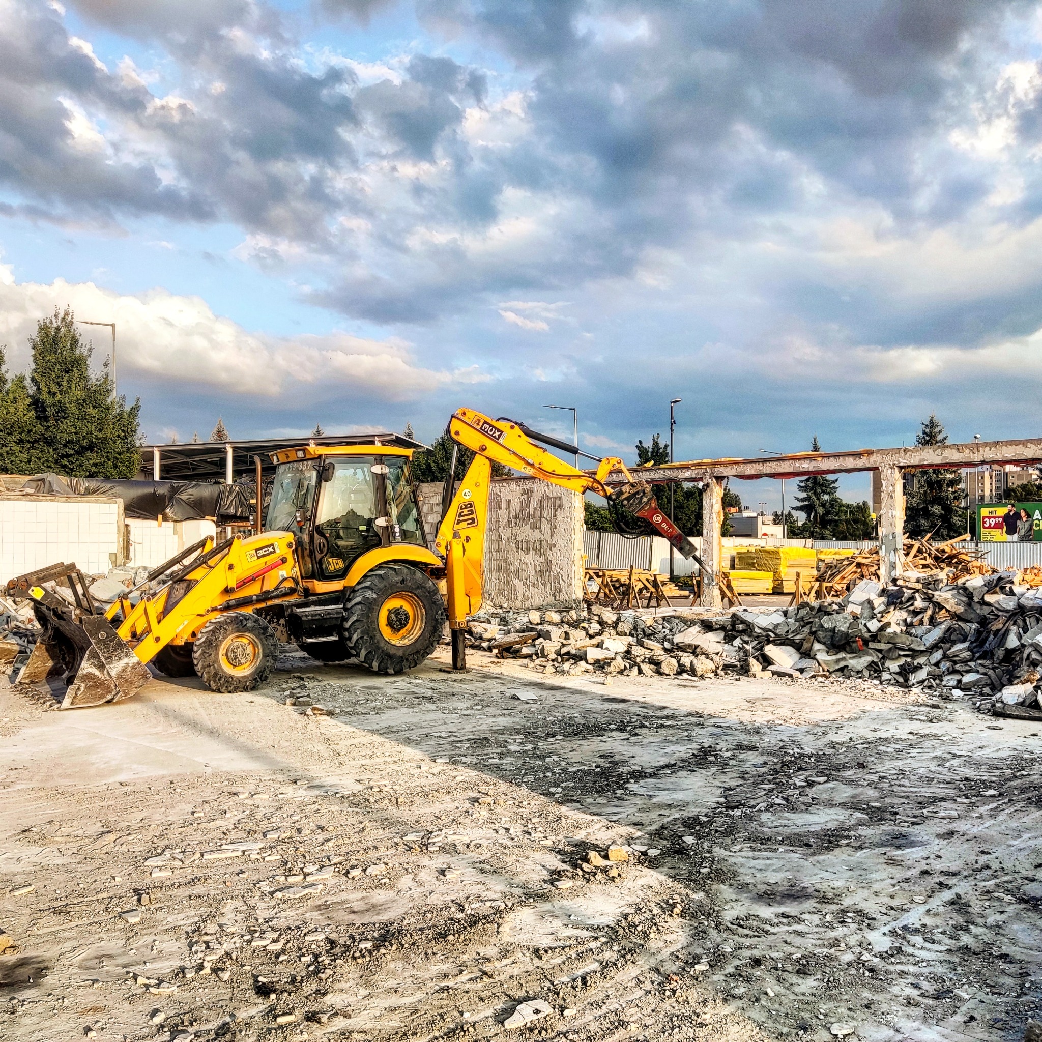 Żółta koparko-ładowarka JCB na placu budowy z gruzem i fragmentami zdemontowanej konstrukcji budynku, w tle widoczne drzewa i pochmurne niebo.