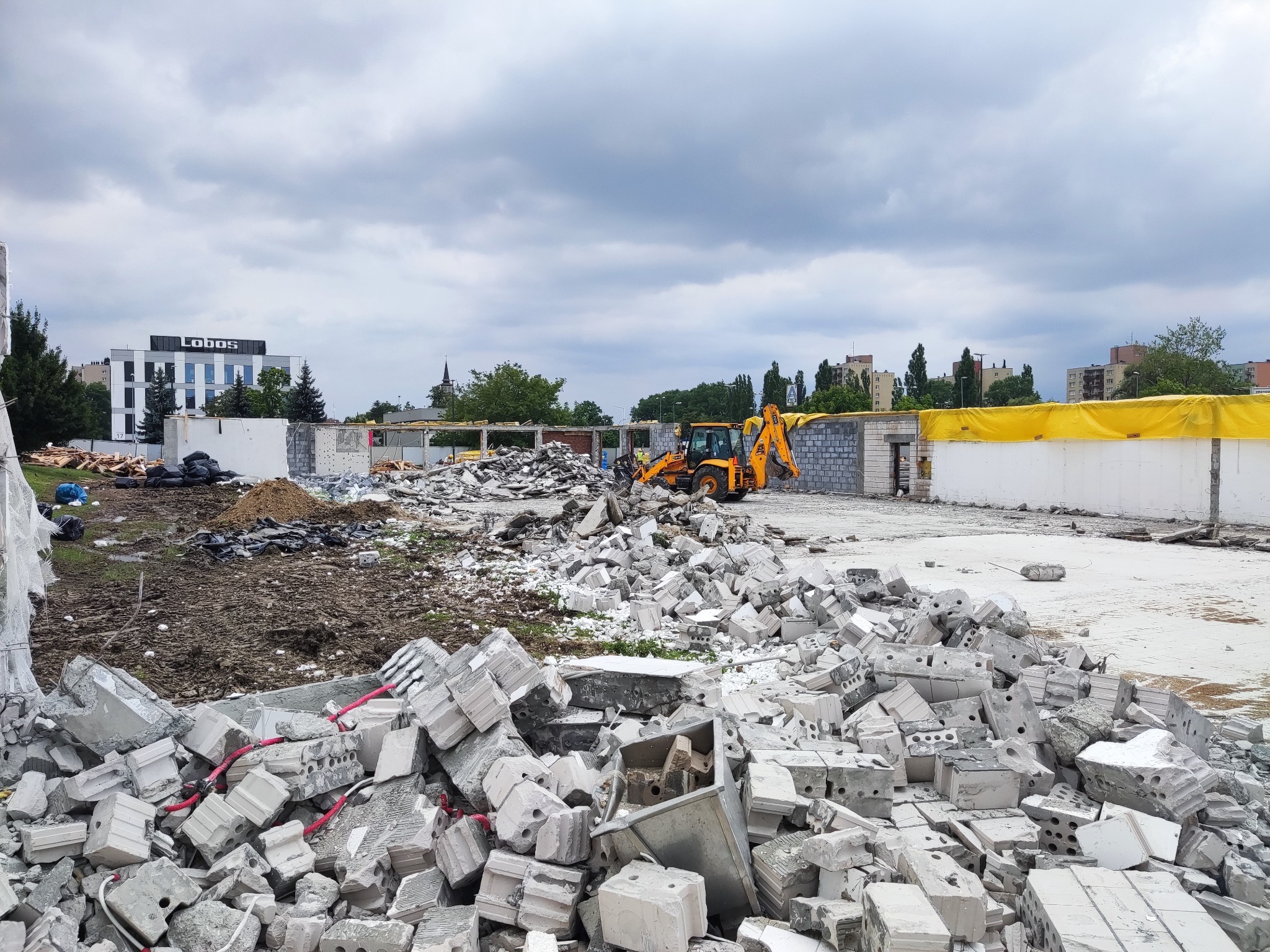 Żółta koparka na placu budowy w Krakowie, wśród gruzu i pozostałości po rozbiórce, z widocznym budynkiem w tle.