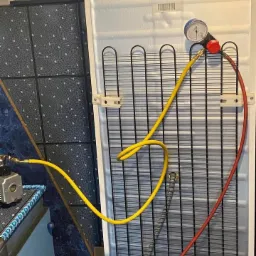 Manometr i przewody podłączone do tylnej kratki chłodnicy lodówki podczas serwisu, widoczna etykieta z informacją o czynniku chłodniczym.