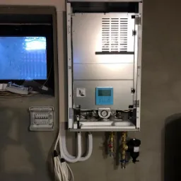 Wnętrze otwartej obudowy urządzenia grzewczego, prawdopodobnie pompy, z widocznymi elementami instalacji, w tym rury, zawory i manometr; na szarej ścianie obok widoczna skrzynka elektryczna i okno.