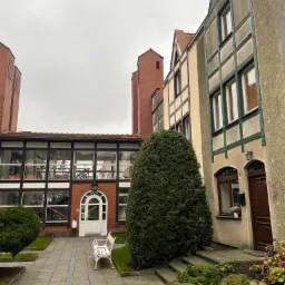 Dziedziniec z nowoczesną architekturą: budynek z szklaną fasadą, biała ławeczka, i budynek mieszkalny z numerem 25, wszystko w stonowanej kolorystyce w pochmurny dzień.