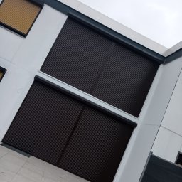 ROMPIS Roman Piszczek - Dwa okna z brązowymi roletami zewnętrznymi, jedno okno z roletą w kolorze złota, zamontowane na białej fasadzie budynku, widok z dołu pod kątem.