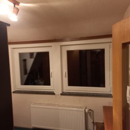 ROMPIS Roman Piszczek - Dwa białe okna w pokoju z grzejnikiem pod nimi. W oknach widoczne odbicia wnętrza. Ujęcie z perspektywy otwartych drzwi.