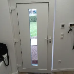 Białe drzwi wejściowe z wąskim przeszkleniem, widok z wnętrza domu na ogród, mata pod drzwiami, widoczne zawiasy i klamka, na podłodze leży narzędzie.