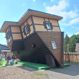 ROMPIS Roman Piszczek - Nietypowy dom z oknami, zbudowany do góry nogami i przechylony pod kątem, z widocznymi drewnianymi elementami konstrukcyjnymi i turystami w tle.