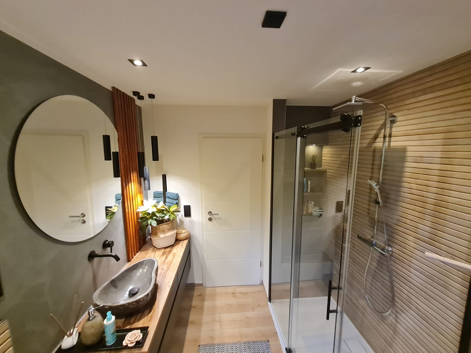 Nowoczesna łazienka z kabiną prysznicową ze szklanymi drzwiami przesuwnymi, umywalką z kamienia na drewnianym blacie i okrągłym lustrem, oświetlona wiszącymi lampami.