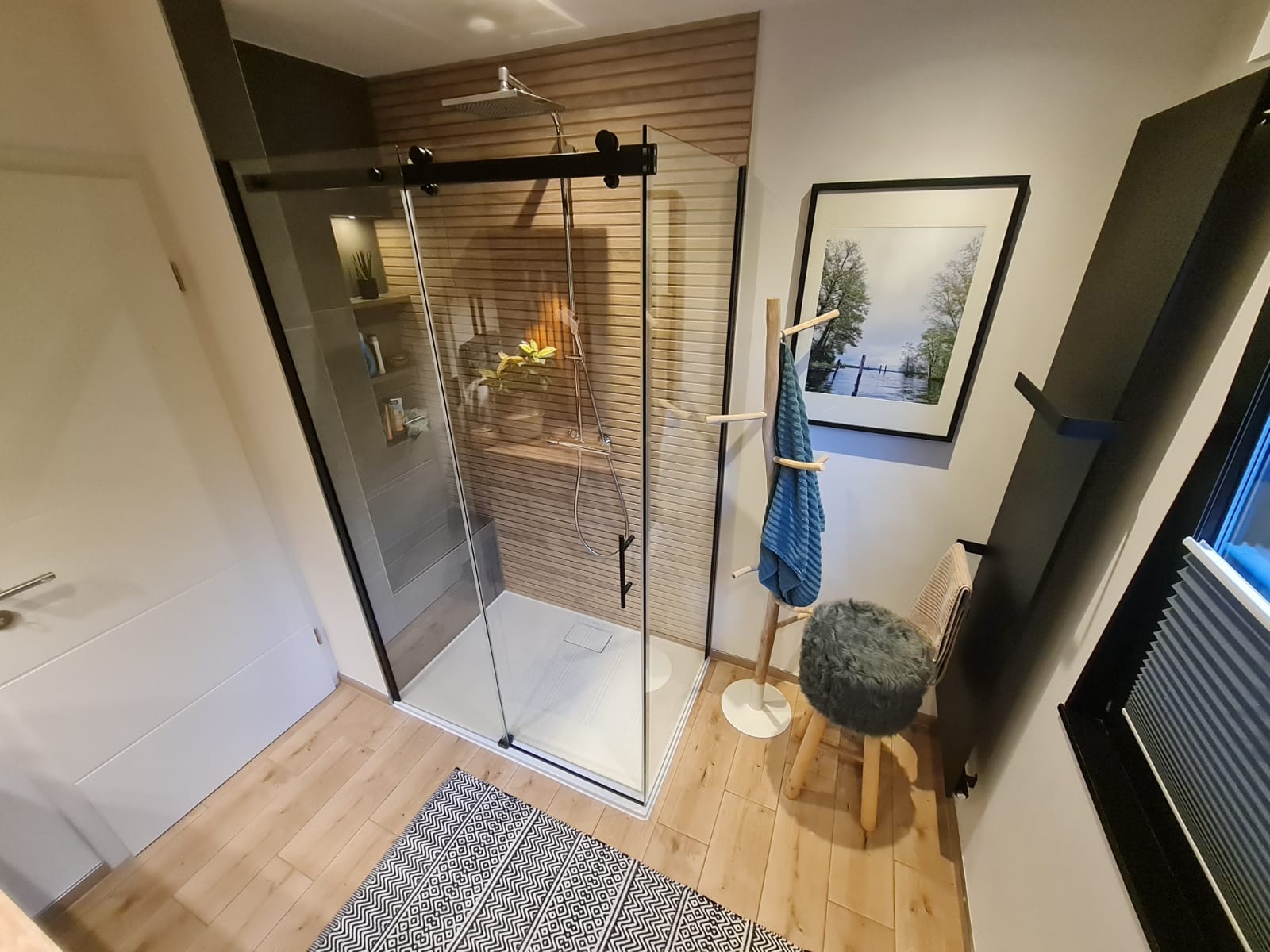 Nowoczesna kabina prysznicowa ze szklanymi przesuwnymi drzwiami na czarnych prowadnicach, drewniana okładzina ściany wewnątrz, widoczna półka z kosmetykami, na zewnątrz drewniana podłoga, dywanik...