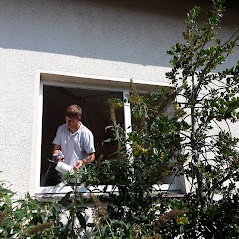 Mężczyzna z silikonem uszczelnia białą ramę okna PCV od wewnątrz, widoczne krzewy na zewnątrz budynku.