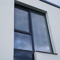 Szare okno z PCV z dwoma skrzydłami o różnej wielkości w białej elewacji budynku, widoczne odbicie nieba.