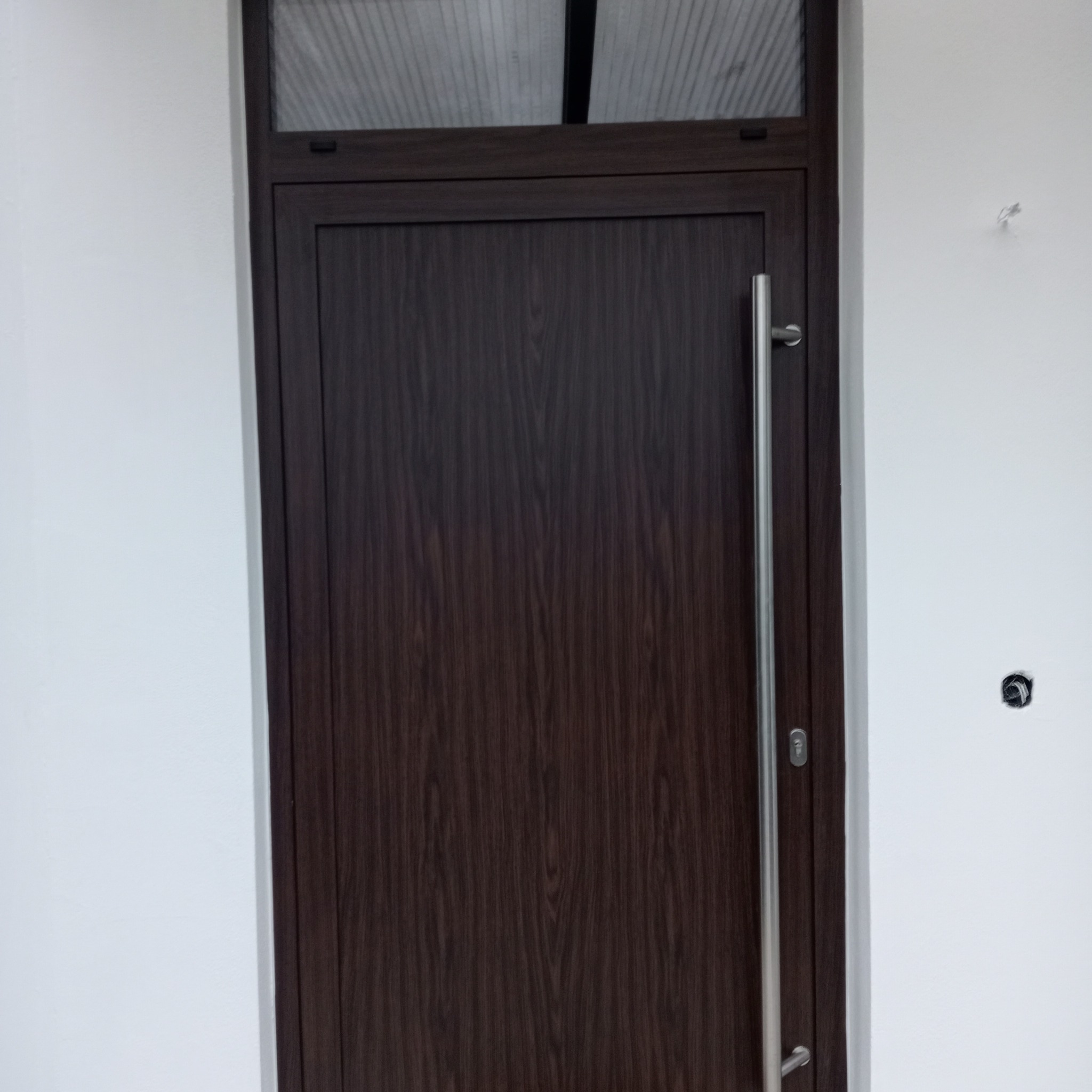 Ciemnobrązowe drzwi wejściowe z drewnopodobnym wykończeniem, długi srebrny uchwyt pionowy, górny naświetlacz z przezroczystego tworzywa, biała ściana z widocznym gniazdkiem elektrycznym bez osprzętu.