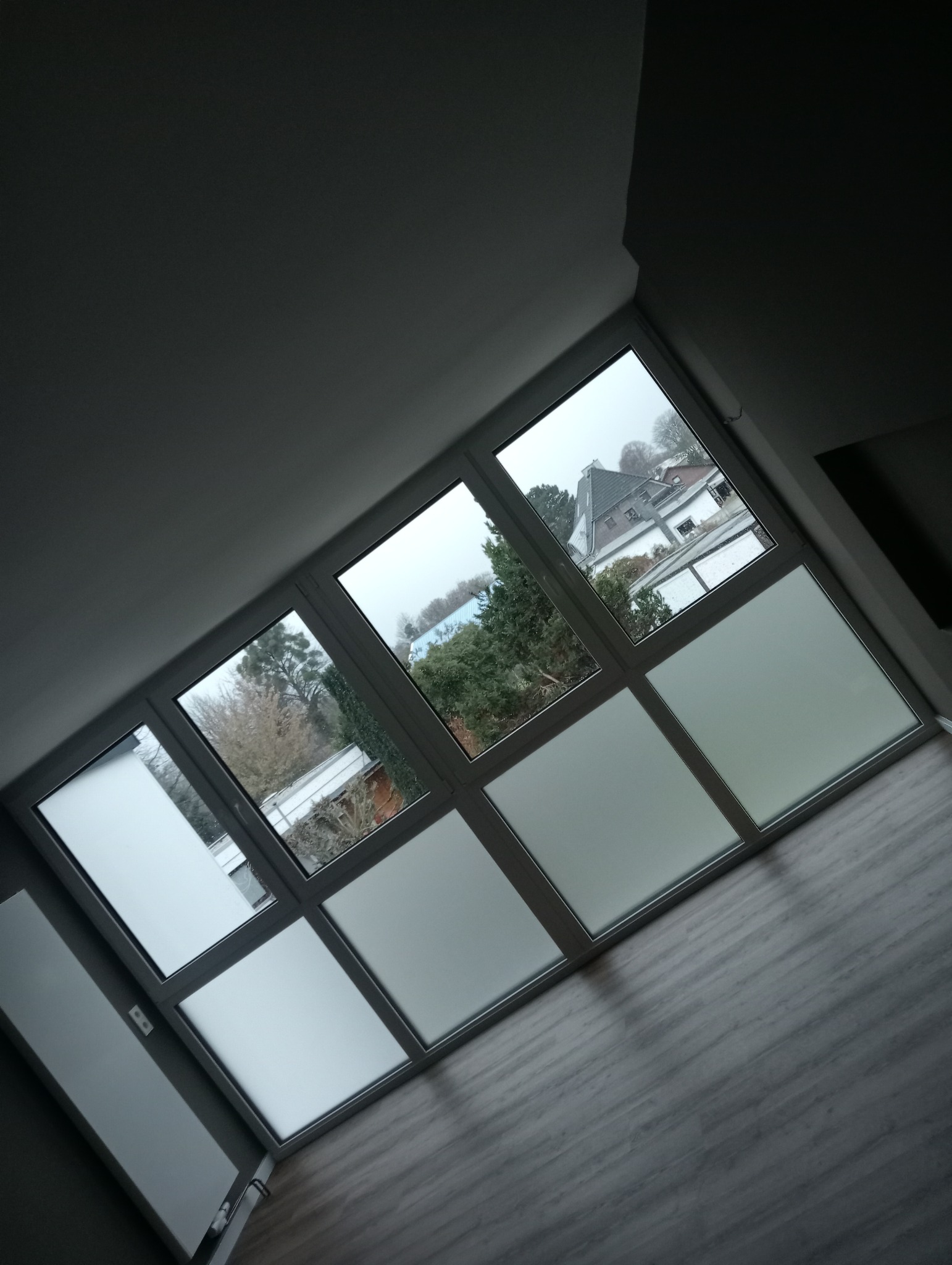 Wnętrze z oknem PCV z matowymi szybami na dolnych panelach, widok na szarą pogodę i domy za oknem, ujęcie z dołu pod kątem.