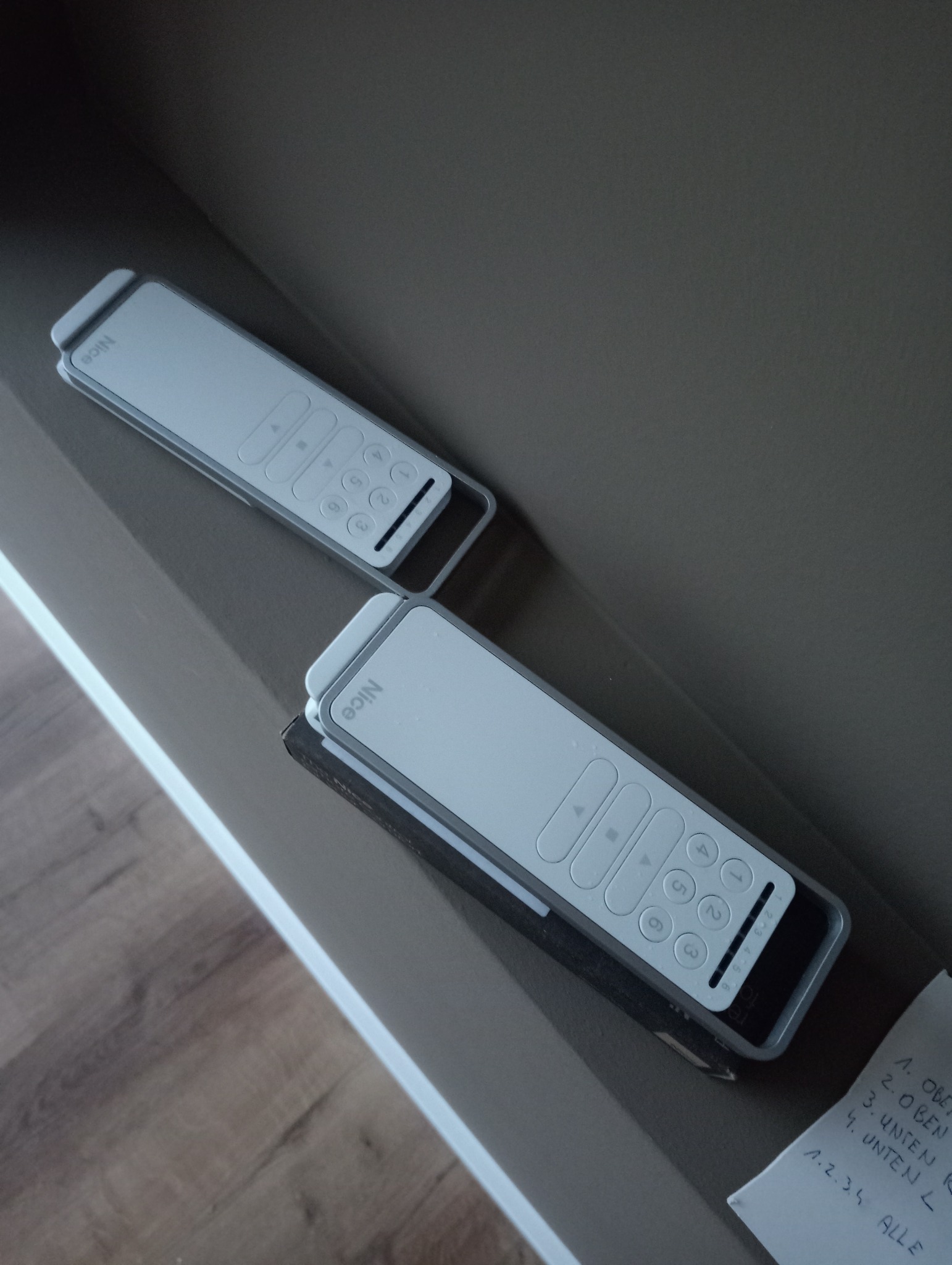 Dwa białe piloty do sterowania roletami leżące na półce, obok kartka z odręcznymi notatkami w języku niemieckim dotyczącymi programowania.