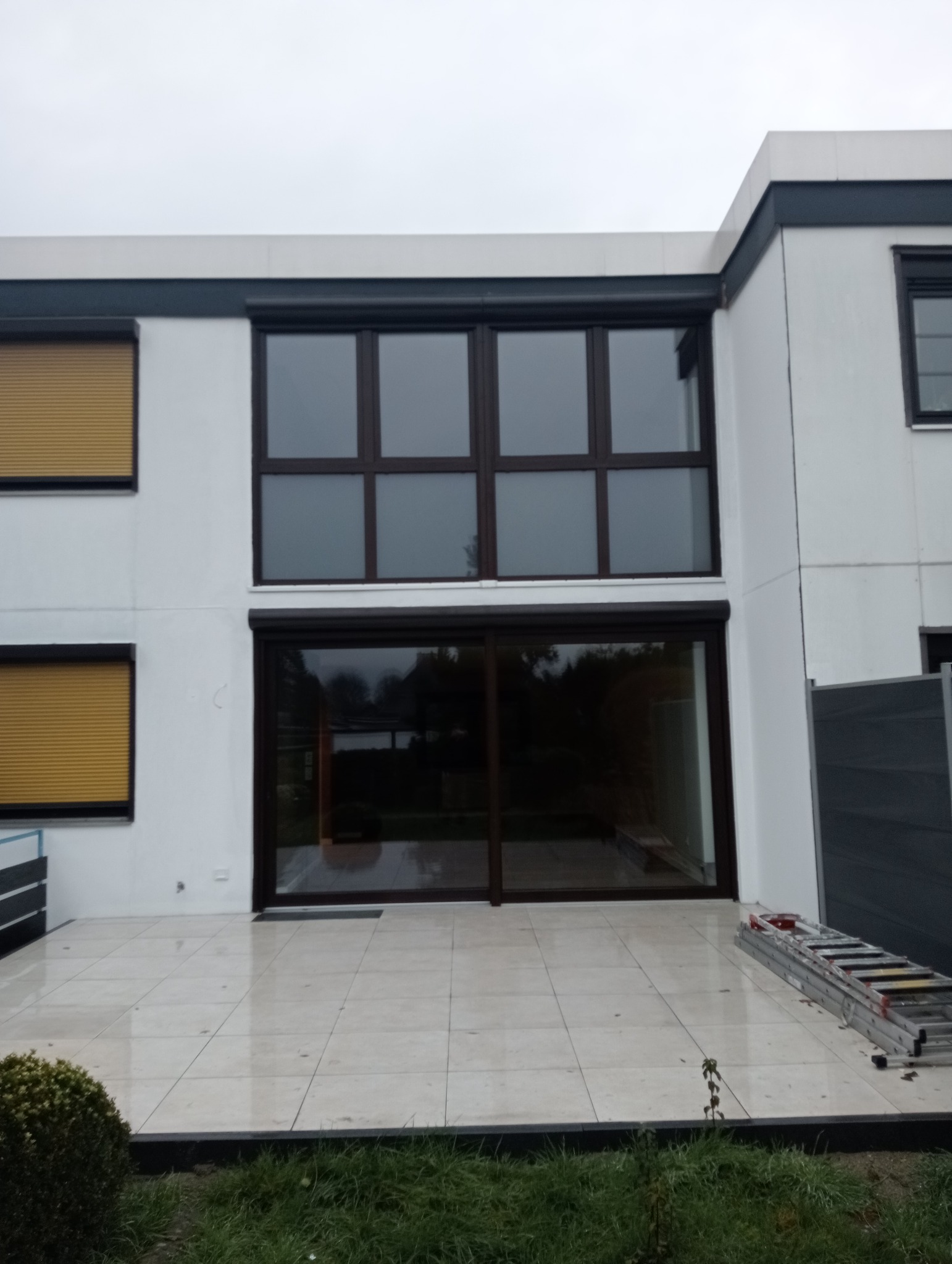 Biały budynek z dużymi przesuwnymi drzwiami szklanymi z ciemną ramą, z roletami zewnętrznymi, widoczna drabina na tarasie z jasnych płytek.