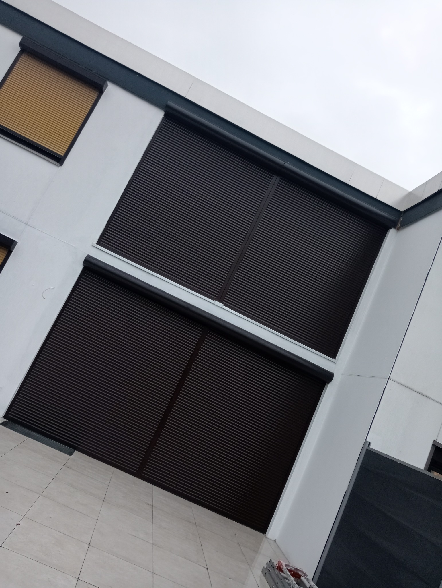 Dwa okna z brązowymi roletami zewnętrznymi, jedno okno z roletą w kolorze złota, zamontowane na białej fasadzie budynku, widok z dołu pod kątem.