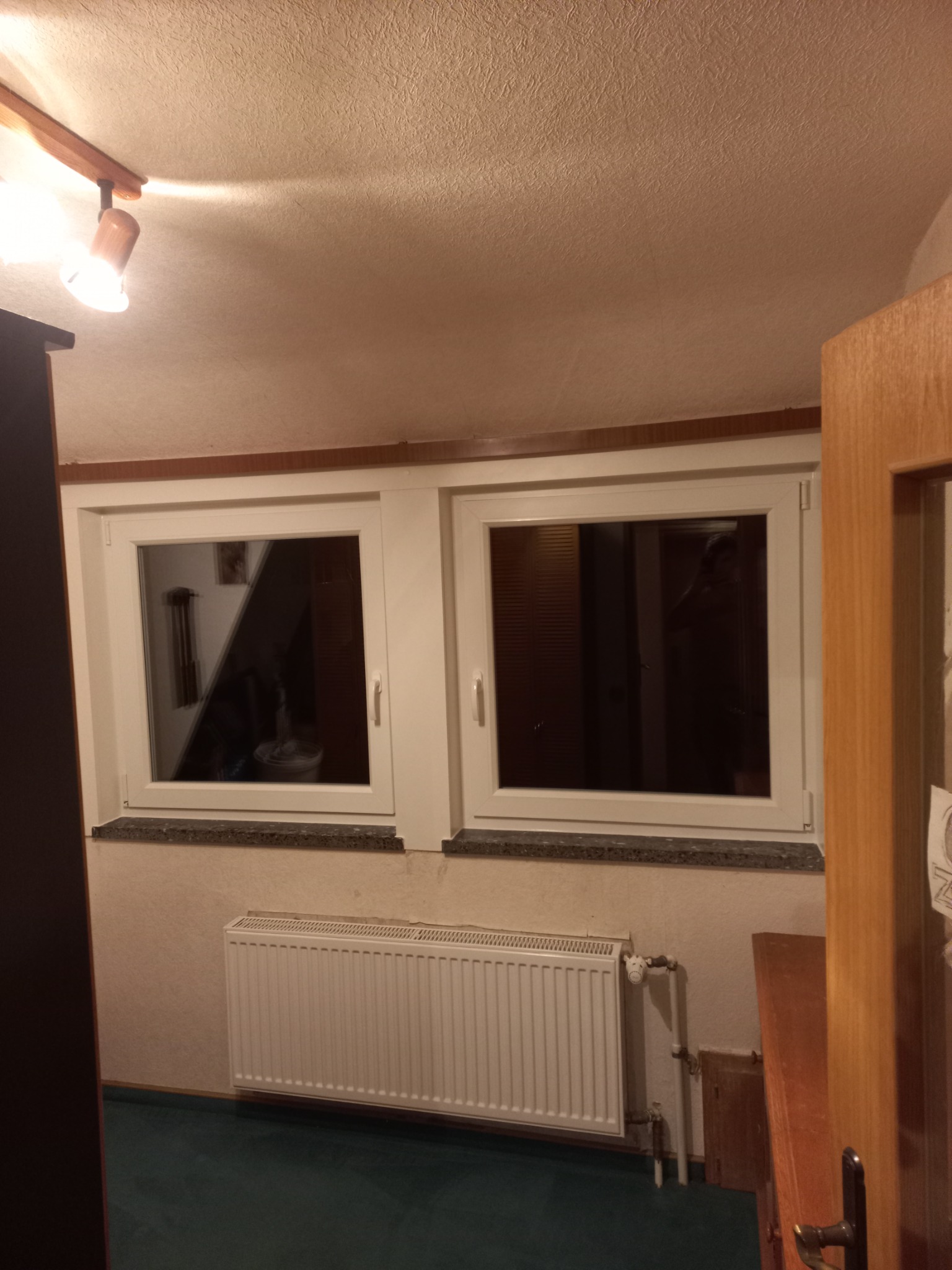 Dwa białe okna w pokoju z grzejnikiem pod nimi. W oknach widoczne odbicia wnętrza. Ujęcie z perspektywy otwartych drzwi.