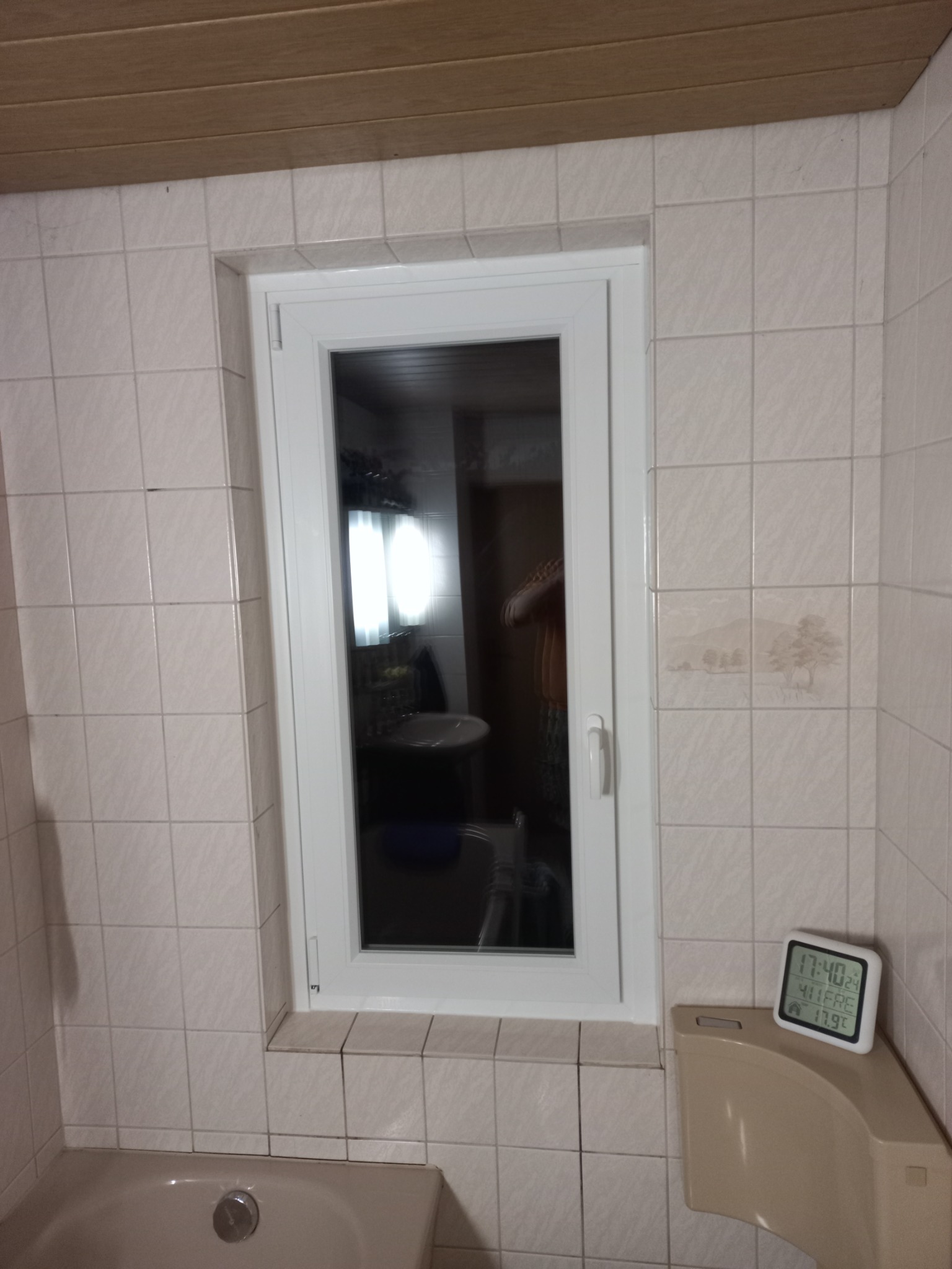 Białe okno PCV w łazience z beżowymi płytkami, widoczny termometr elektroniczny wskazujący godzinę i temperaturę, odbicie wnętrza łazienki w szybie okna.