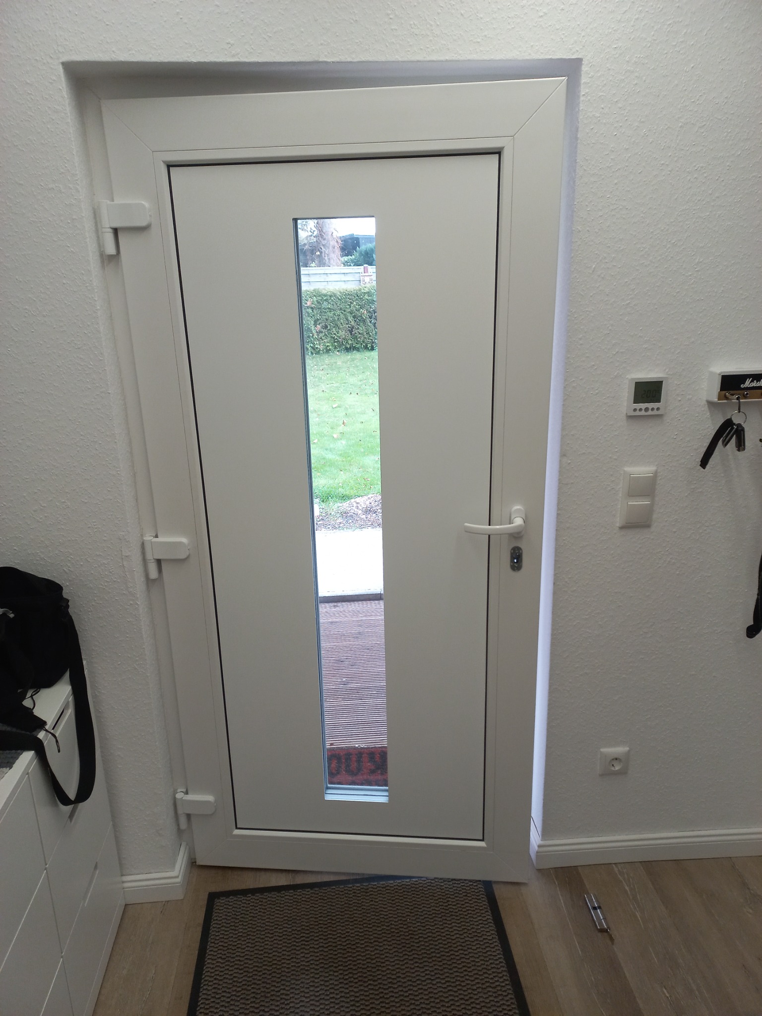 Białe drzwi wejściowe z wąskim przeszkleniem, widok z wnętrza domu na ogród, mata pod drzwiami, widoczne zawiasy i klamka, na podłodze leży narzędzie.