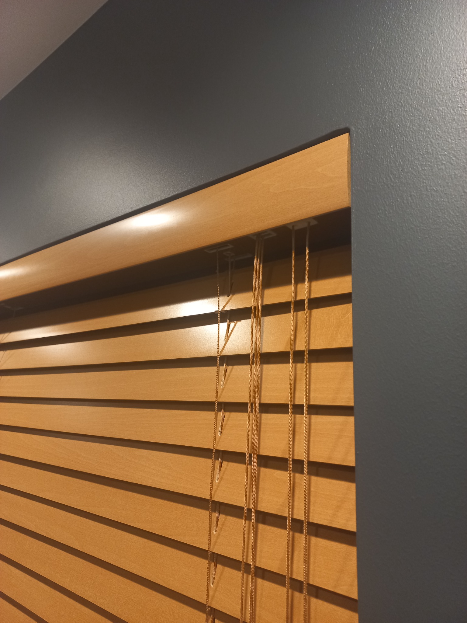 Fragment drewnianych żaluzji poziomej z widocznymi sznurkami sterującymi, na tle szarej ściany.