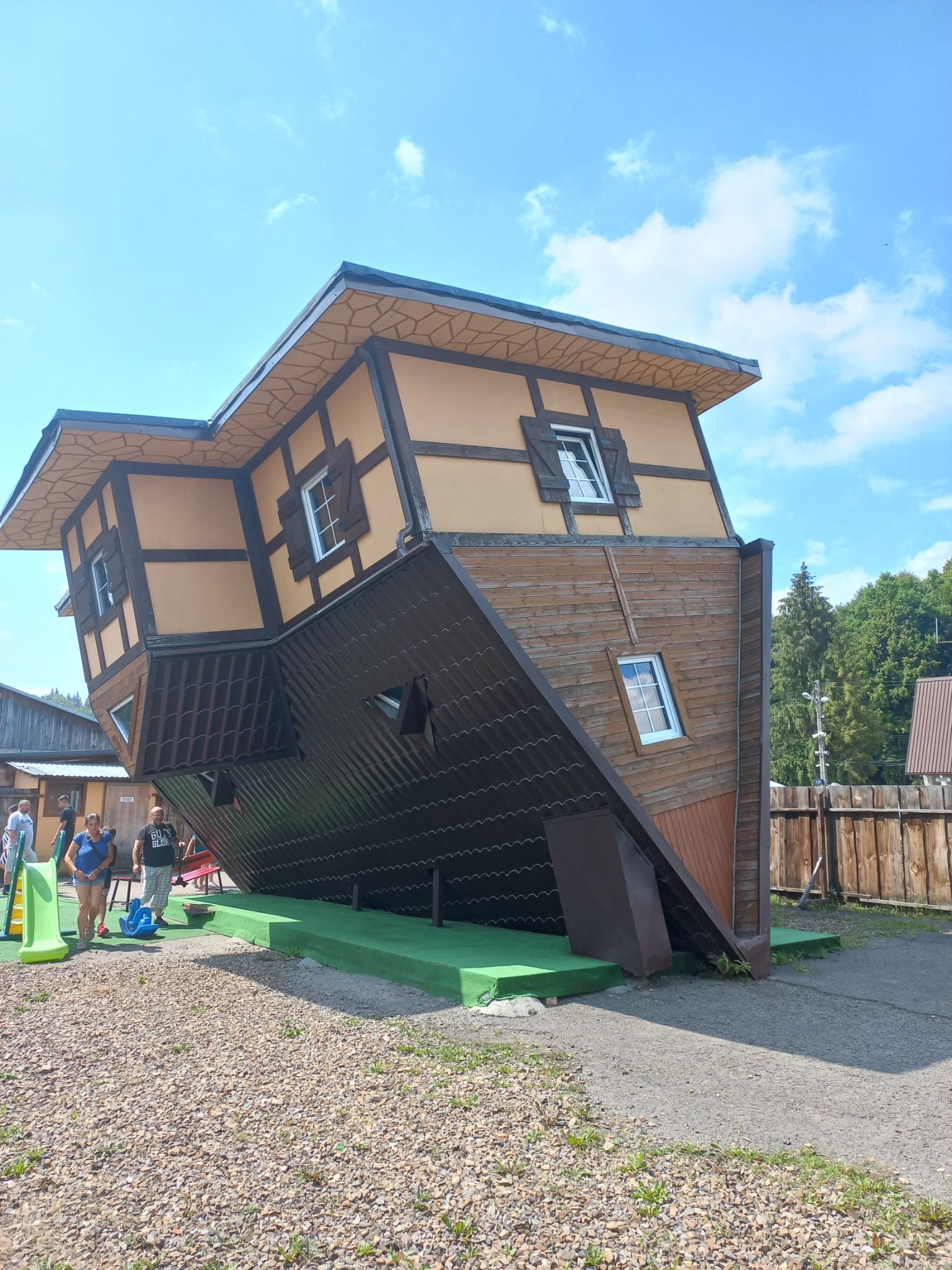 Nietypowy dom z oknami, zbudowany do góry nogami i przechylony pod kątem, z widocznymi drewnianymi elementami konstrukcyjnymi i turystami w tle.