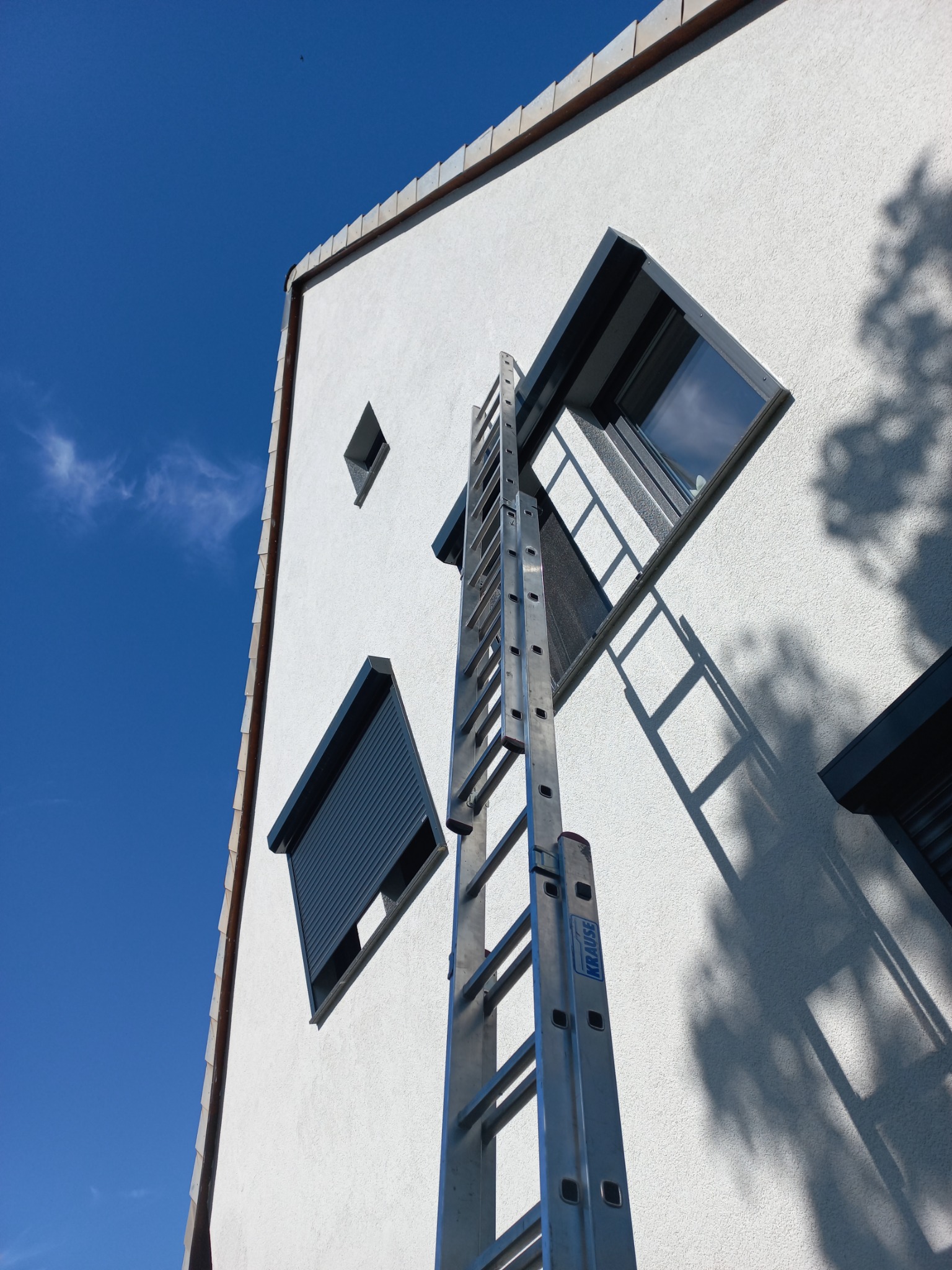 Elewacja budynku z oknami o nietypowym kształcie, jedno z roletą zewnętrzną, drabina oparta o ścianę, błękitne niebo w tle.