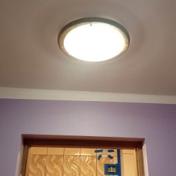 K & J Remont - Fioletowa ściana po malowaniu z kremowym sufitem, oświetleniem sufitowym i białym gzymsem, widok na łazienkę z kafelkami zabezpieczonymi taśmą malarską.