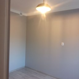 K & J Remont - Pokój po odświeżeniu z szarą farbą na ścianach, jasną podłogą imitującą drewno i nowoczesną lampą sufitową z metalowym kloszem.