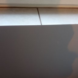 K & J Remont - Fragment ściany pomalowanej na szaro, z widocznymi białymi płytkami u góry i brązową fugą. Ujęcie z bliska, zdominowane przez szarość.