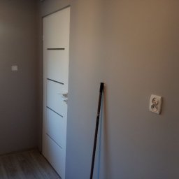K & J Remont - Świeżo pomalowane szare ściany w pokoju z białymi drzwiami z poziomymi wgłębieniami i gniazdkiem elektrycznym, widoczny kij malarski oparty o ścianę.
